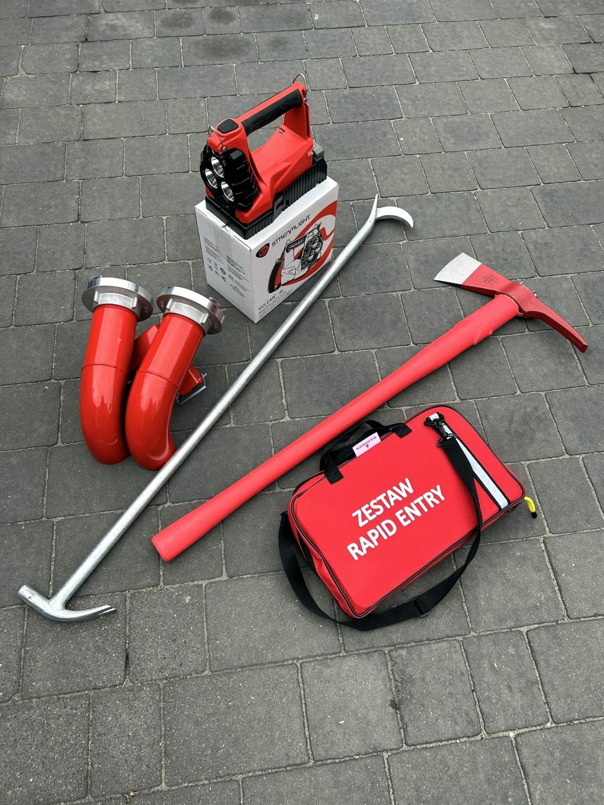 Sprzęt dla OSP Białołęka leży na chodniku : nalewak 2×75, latarka/szperacz Vulcan 180 z ATEX i stacją dokującą od Streamlight, zestaw Rapid Entry od firmy Kadimex, bosak podręczny, topór izolowany.