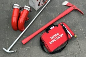 Sprzęt dla OSP Białołęka leży na chodniku : nalewak 2×75, latarka/szperacz Vulcan 180 z ATEX i stacją dokującą od Streamlight, zestaw Rapid Entry od firmy Kadimex, bosak podręczny, topór izolowany.