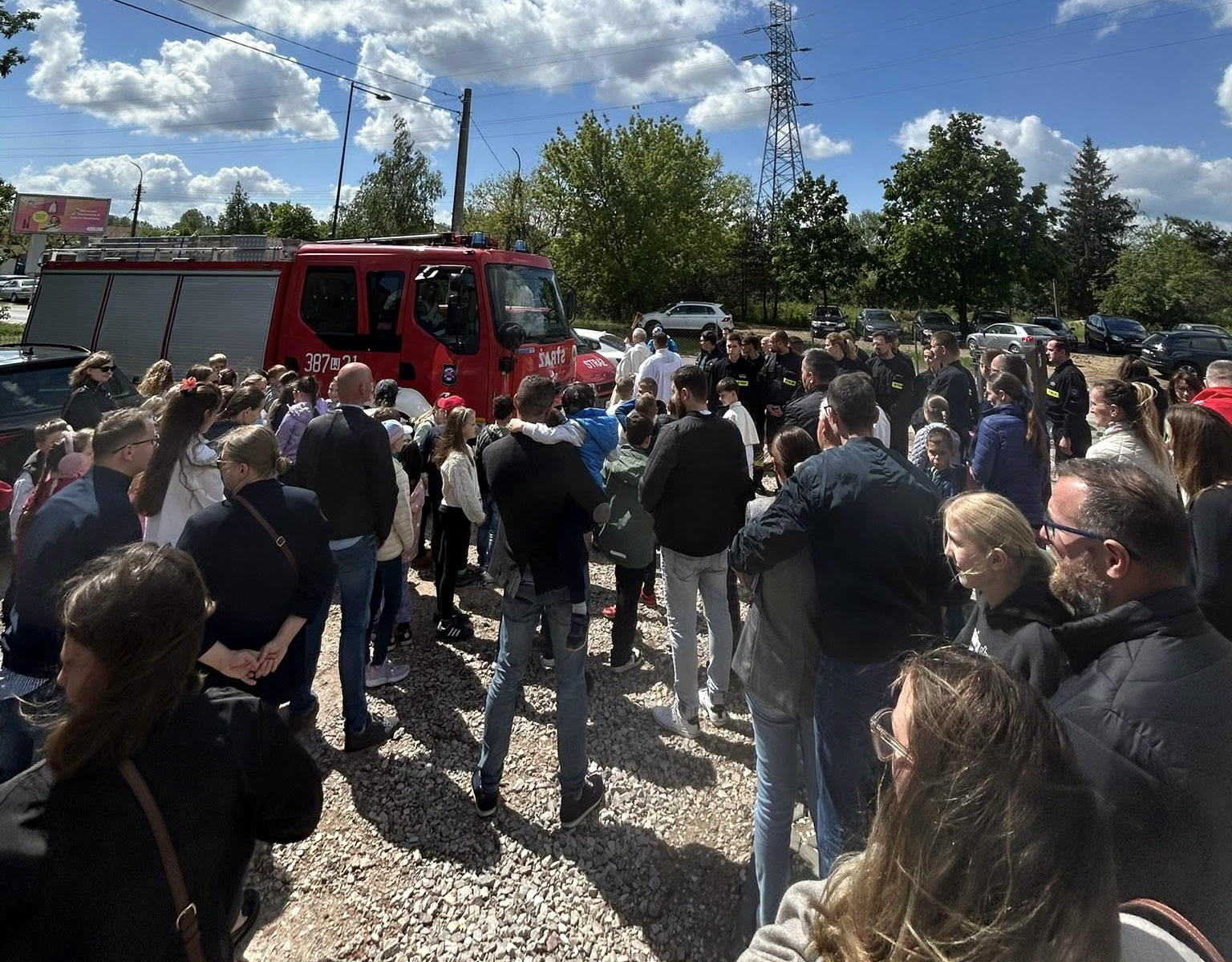 Mieszkańcy zgromadzeni wokół wozu strażackiego podczas obchodów Międzynarodowego Dnia Strażaka, 4 maja. W tle widoczne drzewa, samochody i niebo z lekkim zachmurzeniem — wydarzenie plenerowe promujące bezpieczeństwo i integrację lokalnej społeczności z jednostkami straży pożarnej.