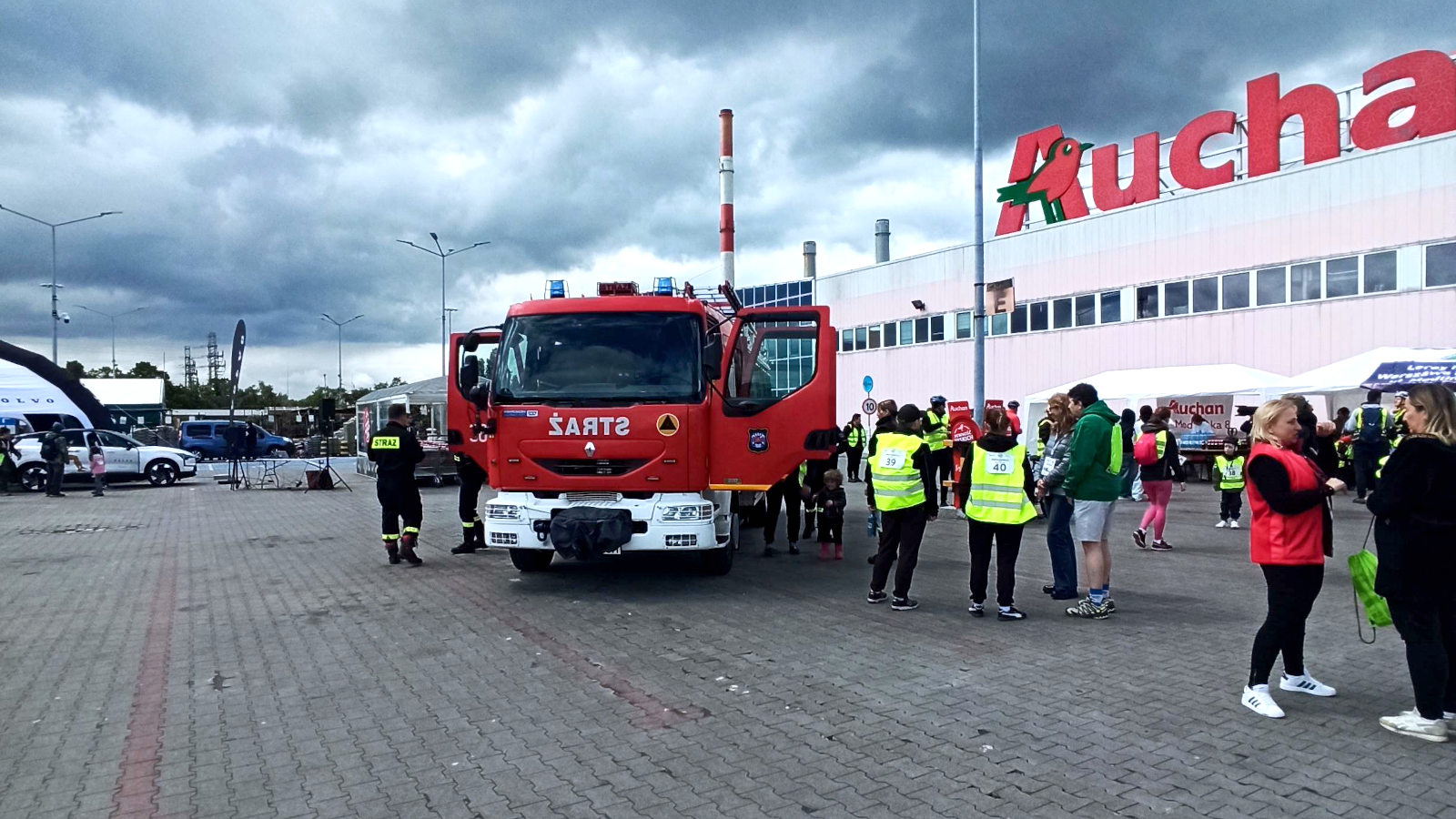 Wóz strażacki OSP Warszawa – Białołęka zaparkowany przed centrum handlowym Auchan podczas wydarzenia towarzyszącego Rodzinnemu Rajdowi Rowerowemu. Strażacy prezentują sprzęt ratowniczy i rozmawiają z mieszkańcami, promując bezpieczeństwo pożarowe i integrując się z lokalną społecznością.